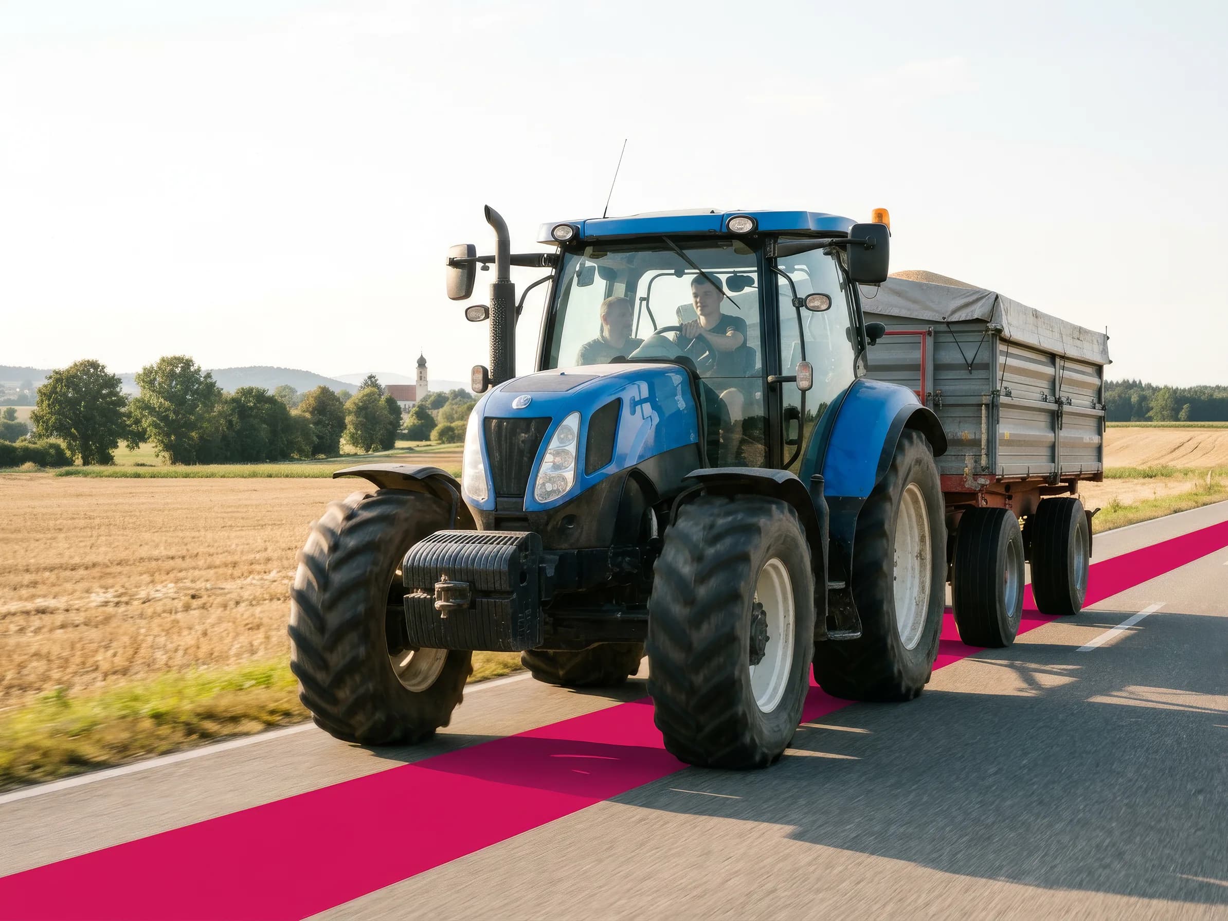 Blauer Traktor mit angehängtem Kipper fährt auf einer Landstraße durch Felder